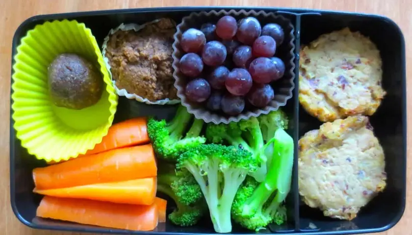 A bento box packed with bento nutrition: a date ball, muffin, grapes, carrots, broccoli, and two savory patties.