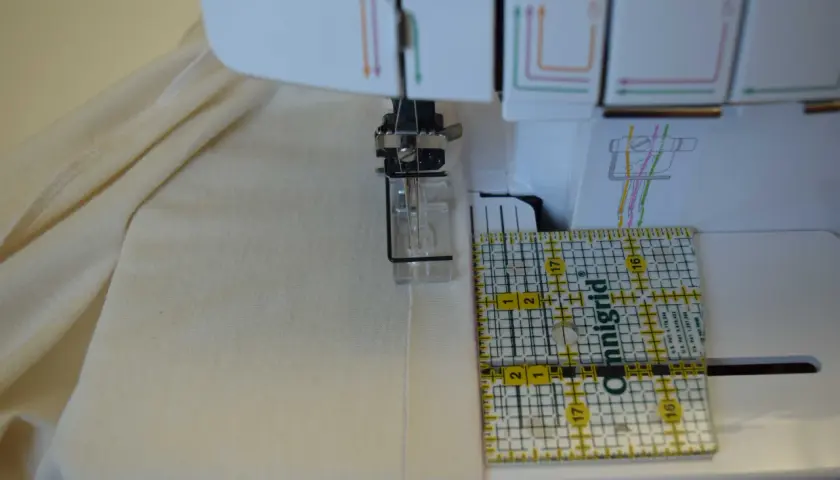 A ruler guides fabric under a coverstitch machine needle, showing how to use a coverstitch machine for precise hemming.