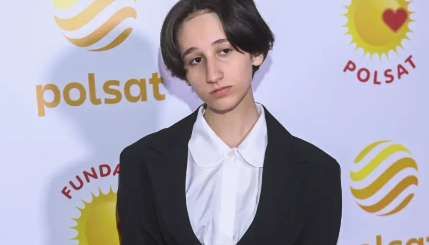 Etiennette Anna Wiśniewska w czarnym żakiecie i białej koszuli z okrągłym kołnierzykiem, na tle logo Polsatu.