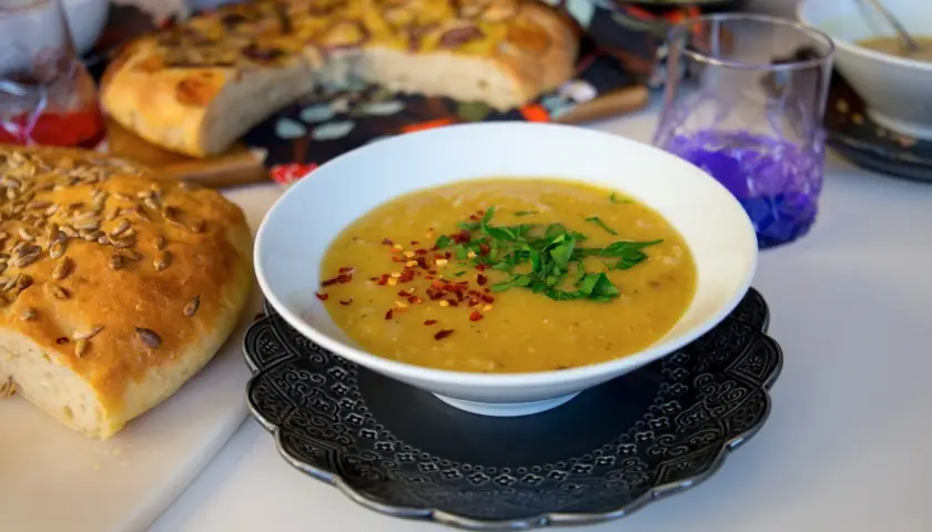 En skål med varm shorbet adas, toppad med persilja och chiliflakes, bredvid ett solrosbröd och focaccia.