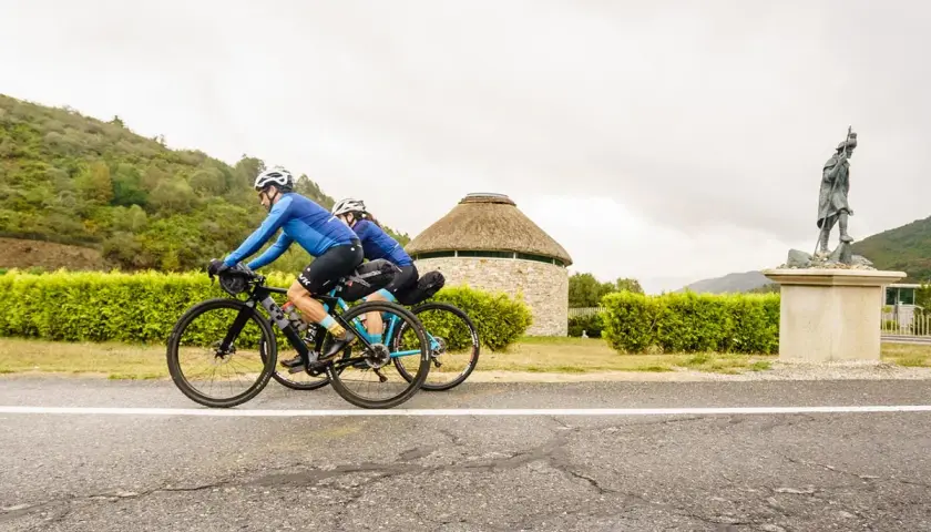 Ciclistas pedalean por la ruta EuroVelo 3, pasando junto a un edificio redondo con techo de paja y una estatua de un peregrino.