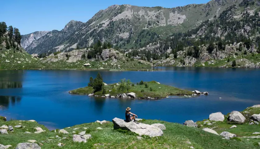 Persona sentada en roca contempla el azul intenso del circ de Colomers, rodeado de montañas verdes y un cielo despejado.
