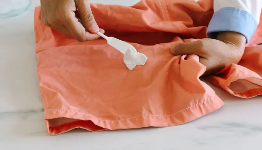 Comment enlever du chewing gum sur un tee shirt : une spatule blanche applique une pâte sur une tache de chewing gum sur un tissu corail.