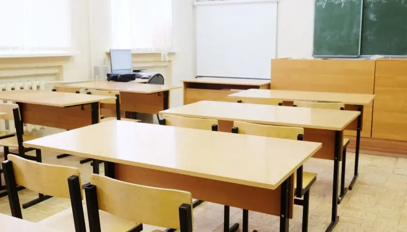 Pusta klasa szkolna z rzędami drewnianych ławek i krzeseł, tablicą i projektorem.
