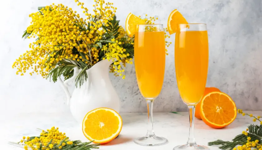 Dwa kieliszki orzeźwiającego drinka mimosa, ozdobione plasterkami pomarańczy, stoją obok bukietu żółtych kwiatów akacji.