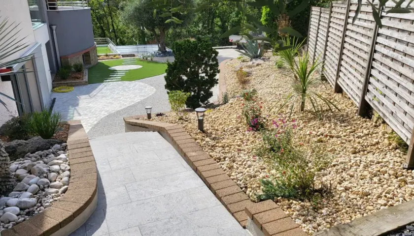 Un aménagement jardin gravier décoratif avec des pas japonais sur pelouse, des plantes et une clôture en bois.
