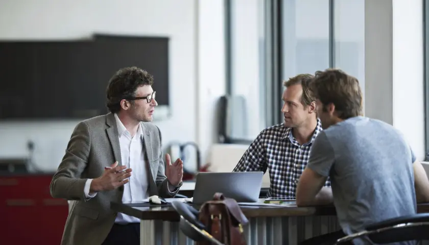 Three men discuss performance review tips for employees in a modern office setting.