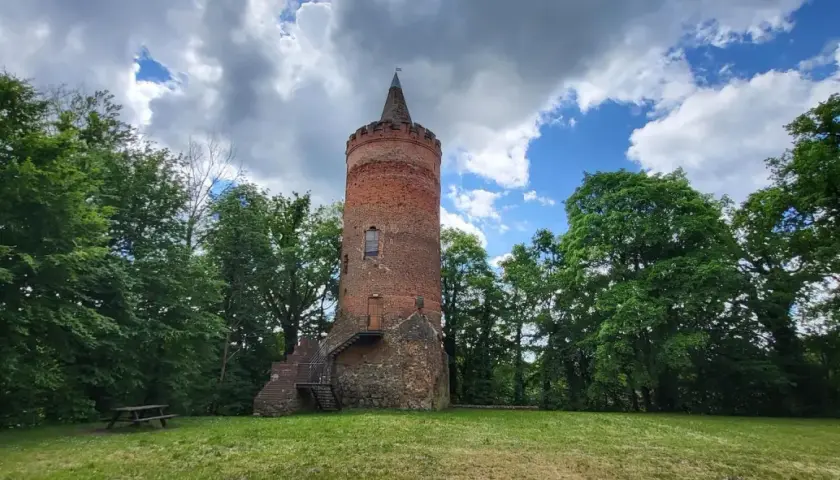 Stara, ceglana wieża zamkowa z kamiennym fundamentem, otoczona zielenią drzew, pod błękitnym niebem z chmurami.