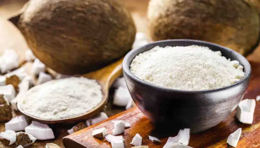 Cocco grattugiato in una ciotola e un cucchiaio, con noci di cocco intere e pezzi di cocco sullo sfondo. Ideale per sostituire la farina di cocco.