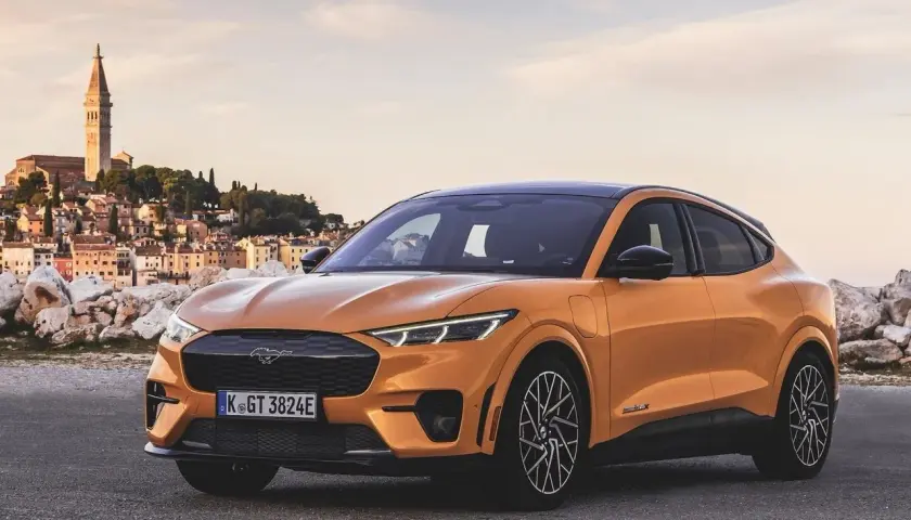 Arancione Ford Mustang Mach-E, una delle auto elettriche sportive più desiderate, parcheggiata con una pittoresca città costiera sullo sfondo.