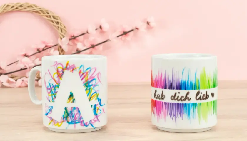 Kreative tassen bemalen ideen: eine Tasse mit dem Buchstaben A und bunte Kritzeleien, daneben eine Tasse mit Regenbogenfarben und der Aufschrift "hab dich lieb".