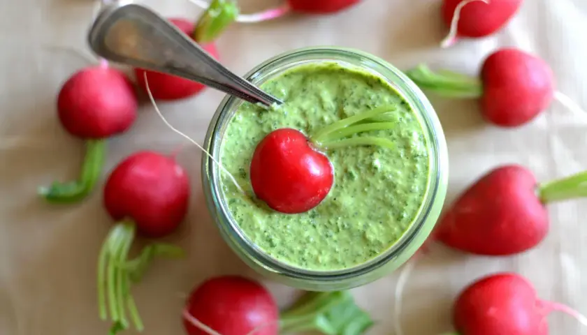 Słoik z zielonym pesto, ozdobiony rzodkiewką i otoczony innymi rzodkiewkami.