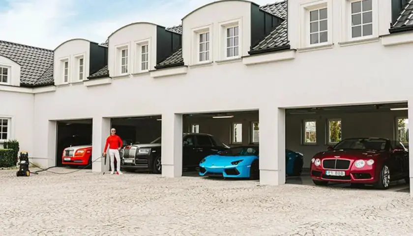 Piotr Śledź w czerwonej bluzie i białych spodniach stoi przy luksusowych samochodach: czerwonym Rolls-Royce, czarnym Rolls-Royce, niebieskim Lamborghini i czerwonym Bentleyu.