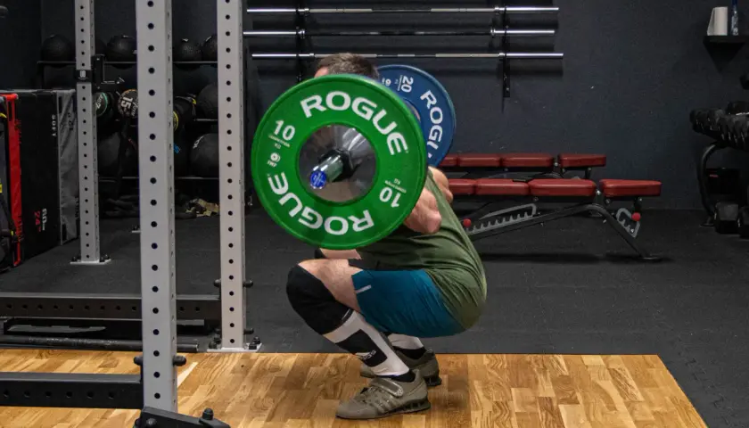 Mężczyzna wykonuje przysiady ze sztangą na karku w siłowni. Zielone talerze "Rogue" ważą 10 kg każdy.