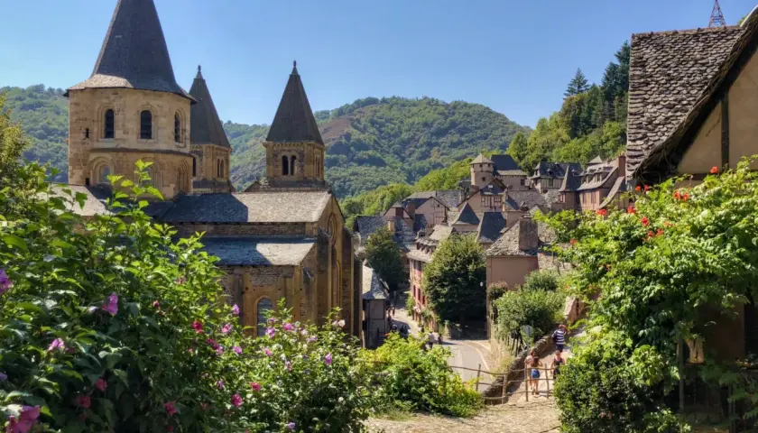 Un pueblo pintoresco con una iglesia románica y tejados de pizarra, ideal para rutas por Francia. Flores rosas y verdes exuberantes en primer plano.