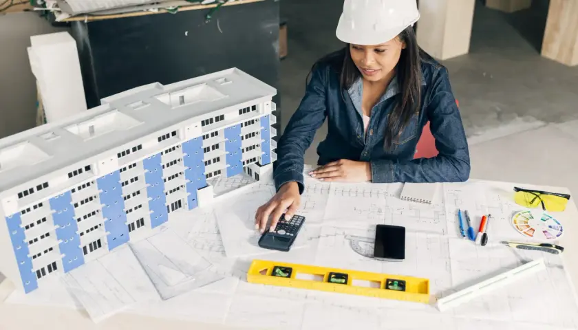 Architektka w kasku nadzoruje model budynku, korzystając z kalkulatora i planów.