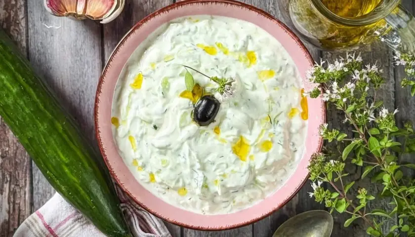 Schüssel mit cremigem griechischem Tzatziki, garniert mit Olive und Öl. Daneben Gurke, Knoblauch und frische Kräuter.