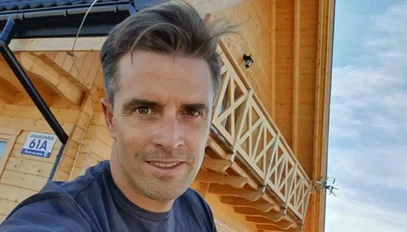 Staszek Karpiel-Bułecka pozuje na tle drewnianego domu z balustradą.