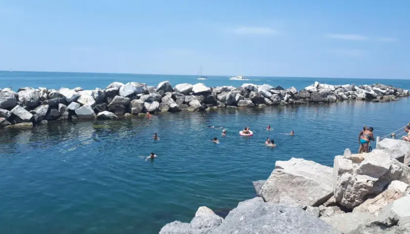 Baignade estivale dans la marina de Pise, avec des rochers formant une digue et des baigneurs profitant du soleil.