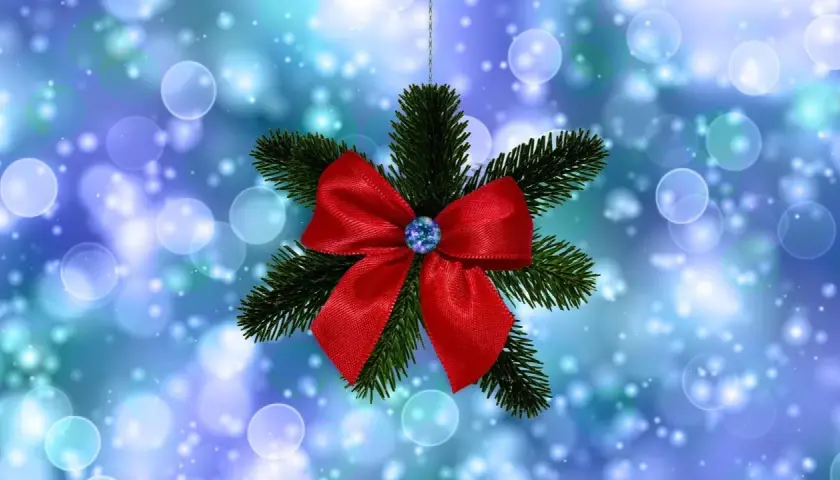 Un lazo de navidad rojo brillante adorna una rama de pino, suspendido sobre un fondo azul con luces bokeh.