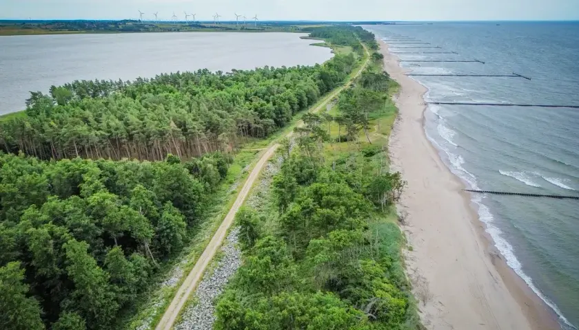 Malownicza nadmorska trasa rowerowa biegnie wzdłuż lasu i plaży, z widokiem na morze i jezioro.