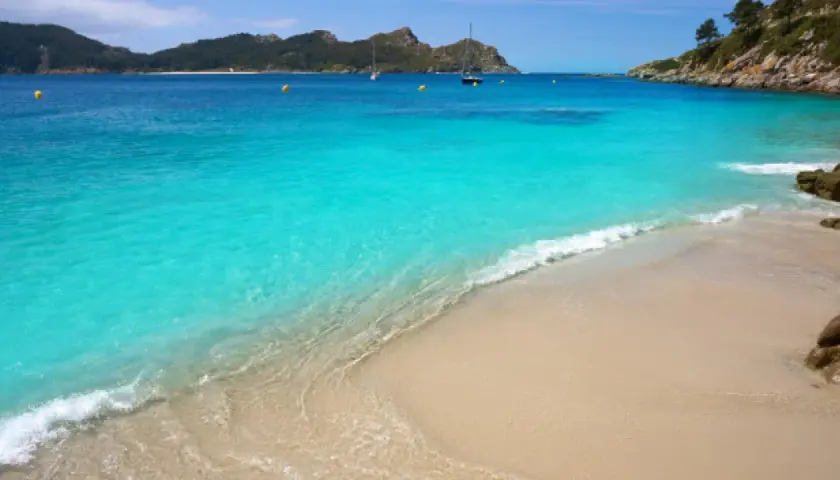 Aguas turquesas y arena dorada en unas playas cristalinas España, con veleros navegando al fondo y montañas verdes.