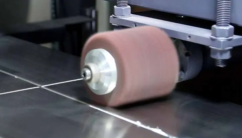 Máquina puliendo acero inoxidable. Un rodillo abrasivo gira sobre una superficie metálica, dejándola brillante.