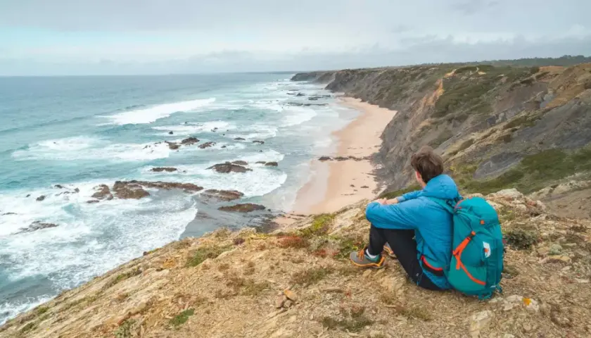 Persona con mochila azul contempla la costa salvaje durante un senderismo Portugal. Olas rompen en la playa desierta.