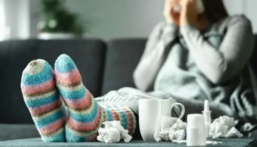 Osoba w kolorowych skarpetkach chora, otulona kocem, z chusteczkami i lekami. Objawy alergii dają się we znaki.