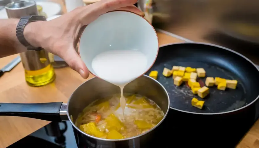 Dodawanie mleczka kokosowego do zupy, by zagęścić ją bez mąki. Na patelni smażą się kostki tofu.