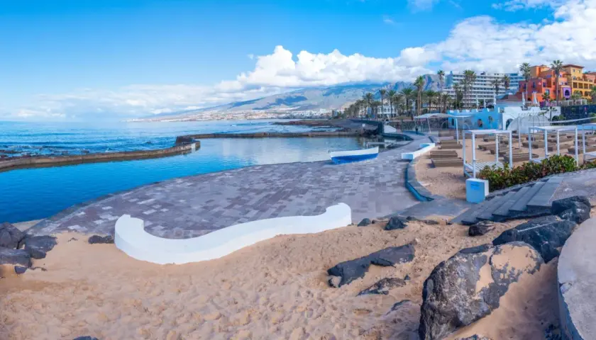 Słoneczna plaża Playa de las Américas z basenem, kolorowymi budynkami i górami w tle.