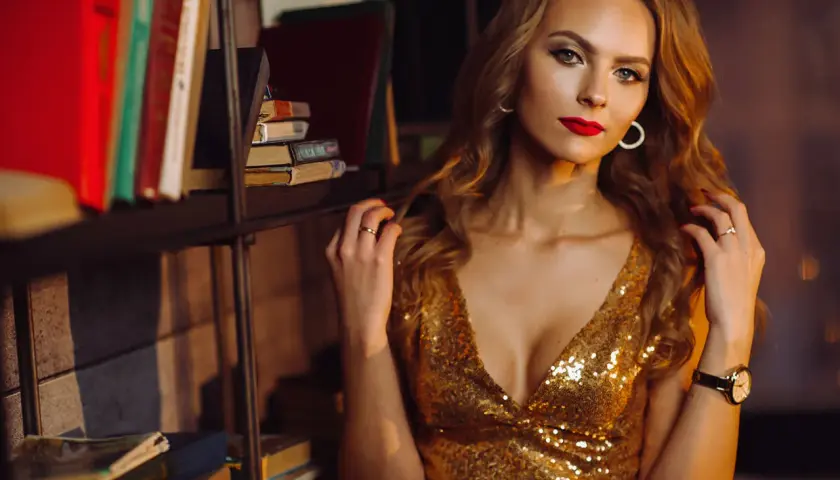 Elegancka kobieta w złotej sukience, z wyrazistym makijażem i czerwoną szminką, pozuje na tle regału z książkami.
