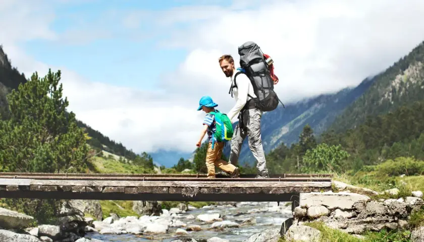 Vater und Sohn überqueren eine Brücke beim Wandern mit Kleinkind. Berge und ein Fluss im Hintergrund.