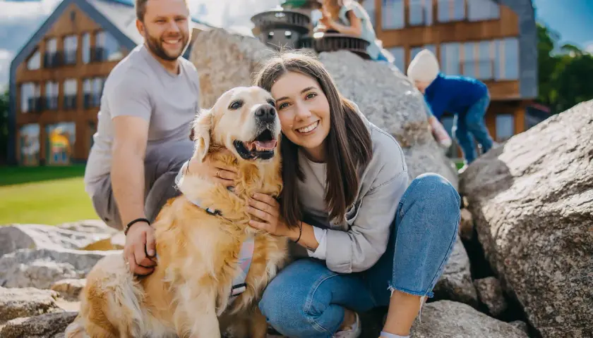 Uśmiechnięta rodzina z psem na tle drewnianych domków. Czy na Śnieżkę można z psem? Tak, jeśli jest tak szczęśliwy jak ten złoty retriever!