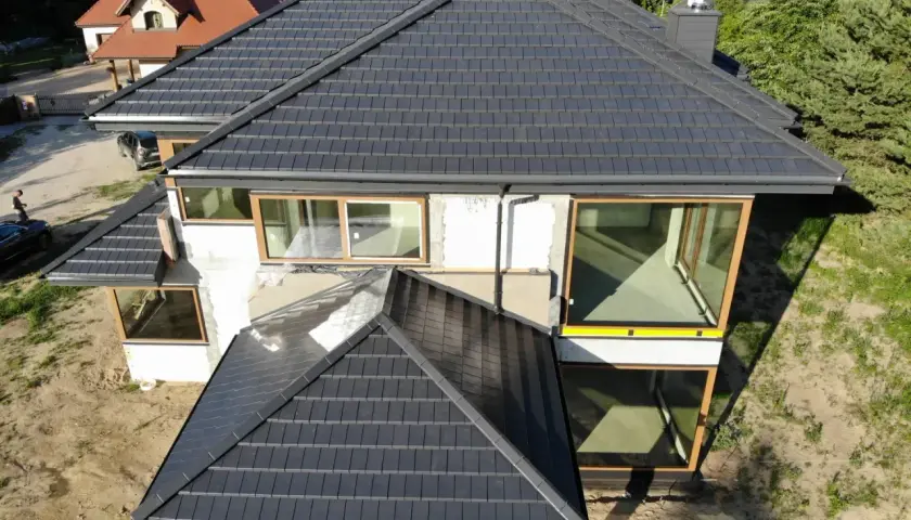 Nowy dom z ciemną dachówką, która wygląda jak blacha. Duże okna z drewnianymi ramami.
