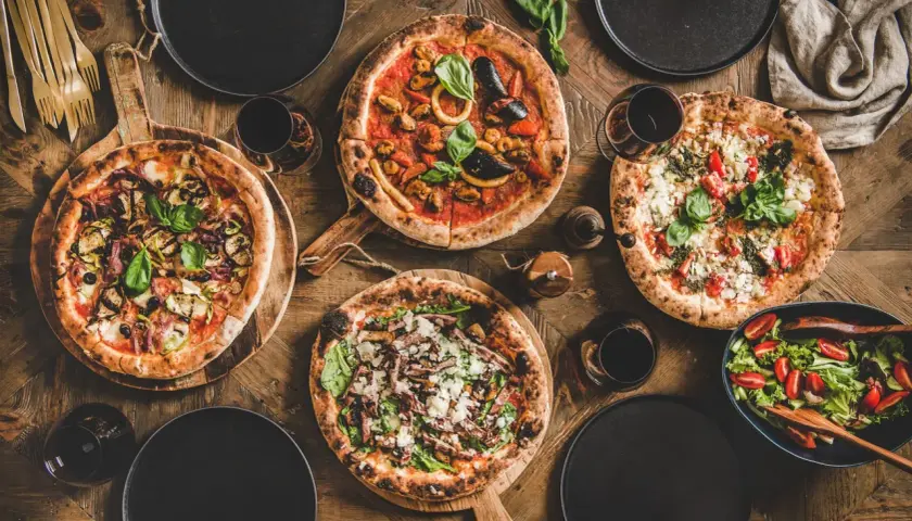 Vier verschiedene Pizzen, ein Salat und Rotwein auf Holztisch. Die Frage welcher Wein zu Pizza wird hier stilvoll beantwortet.