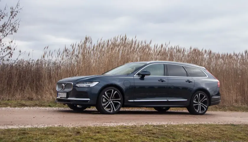 Ciemnogranatowe Volvo v90 na tle wysokiej, suchej trawy i pochmurnego nieba.