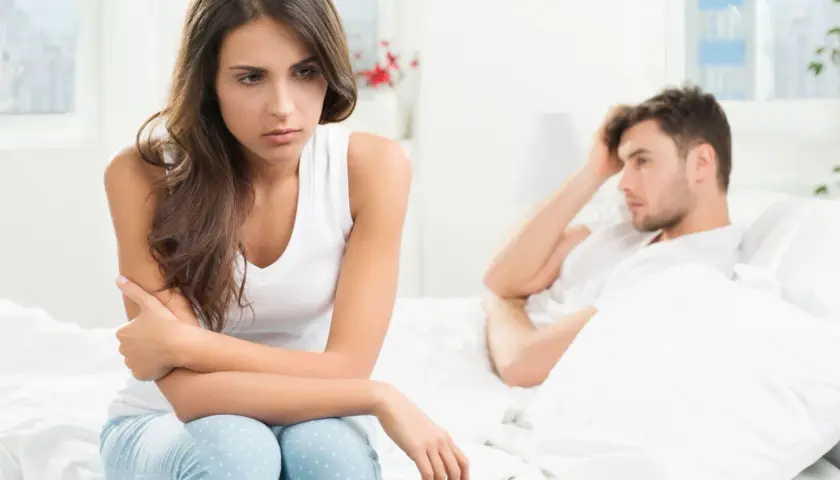 Smutna kobieta siedzi na łóżku, zmartwiona. Jej partner leży obok, z ręką na czole. Czy to uczulenie na spermę powoduje ich problemy?