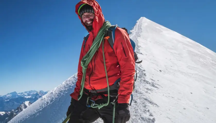 Uśmiechnięty alpinista w czerwonej kurtce na szczycie góry, z liną na ramieniu, na tle majestatycznego **Mount Blanc**.