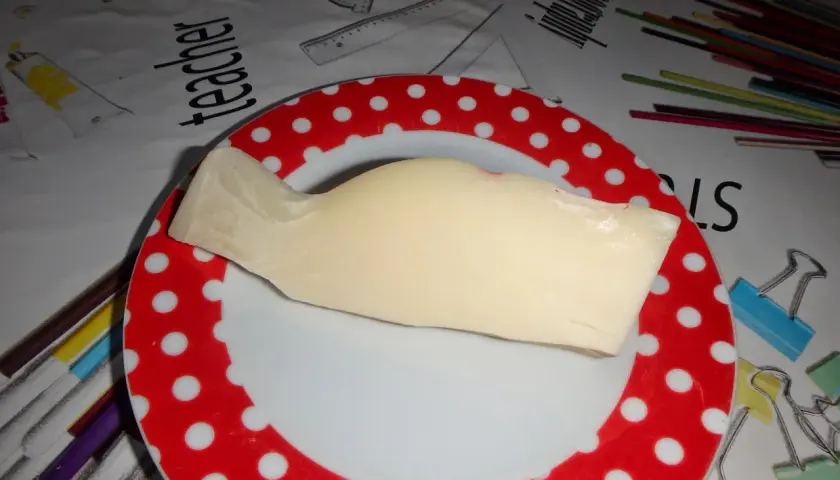 Kawałek żółtego sera provolone leży na talerzu w czerwone kropki. Tło przedstawia materiał z rysunkami przyborów szkolnych.