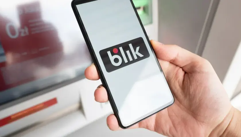 Dłoń trzyma smartfon z logo Blik przed bankomatem. Uważaj na oszustów, którzy mogą próbować wyłudzić dane.