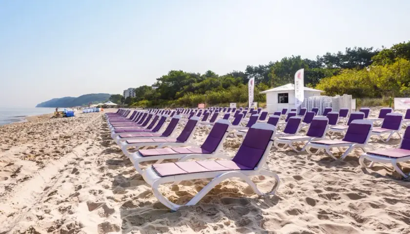 Rzędy leżaków na piaszczystej plaży, idealne na tygodniowe wczasy nad morzem z wyżywieniem. W tle morze i zieleń.