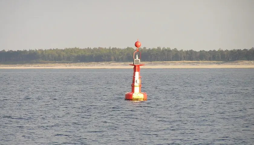 Czerwono-biała boja nawigacyjna, znak bezpiecznej wody, na spokojnym morzu. W tle linia lasu i plaży.
