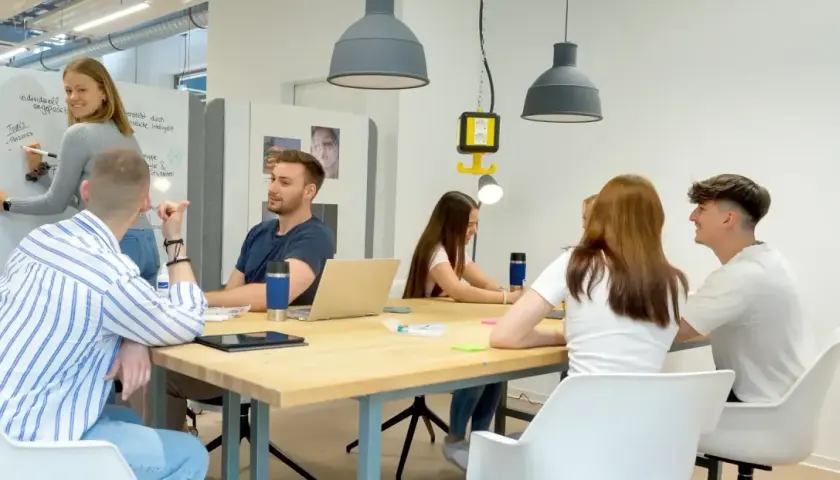 Junge Leute im **Master berufsbegleitend NRW** diskutieren an einem Tisch. Eine Frau schreibt an einer Tafel.