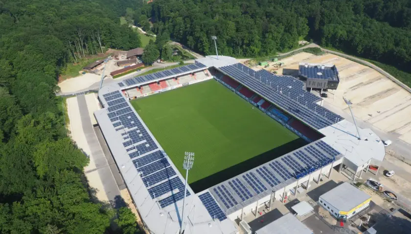 Widok z lotu ptaka na zieloną murawę i trybuny stadionu, otoczonego lasem. To najmniejszy stadion na świecie, z panelami słonecznymi na dachu.