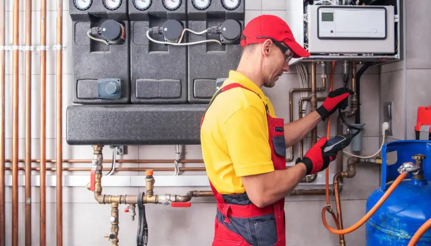 Instalator w czerwonej czapce i żółtej koszulce wykonuje próbę szczelności gazu, używając detektora przy instalacji grzewczej i butli gazowej.
