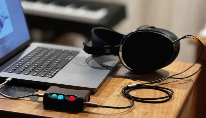 Ein Laptop, ein Kopfhörerverstärker mit leuchtenden LEDs und ein Kopfhörer mit großem **headphone driver** liegen auf einem Holztisch.