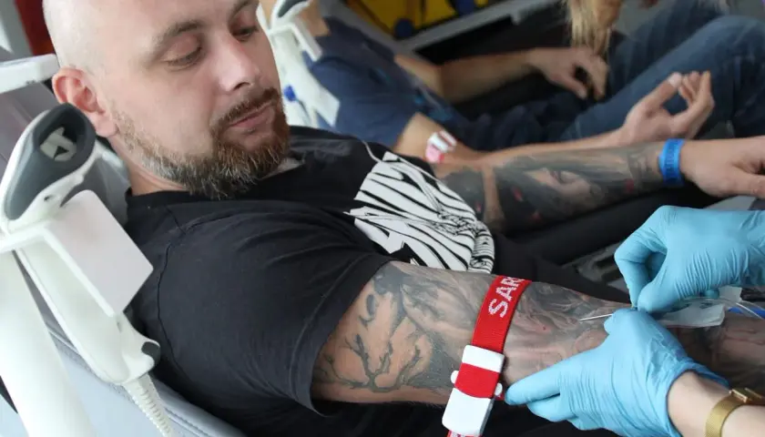 Mężczyzna z tatuażem na ramieniu oddaje krew. Czerwona opaska uciskowa i igła są widoczne.