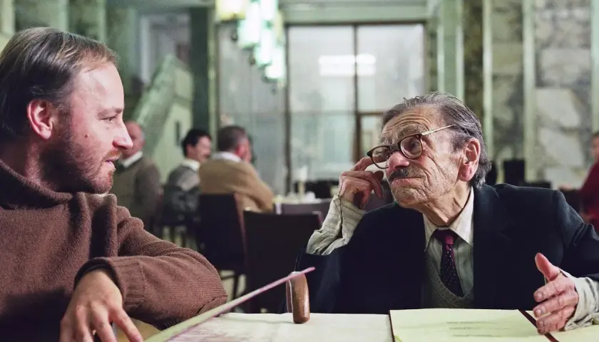 Dwóch mężczyzn rozmawia przy stole. Jeden z nich, starszy, w okularach, gestykuluje. Scena z jednego z filmów Krzysztofa Krauzego.