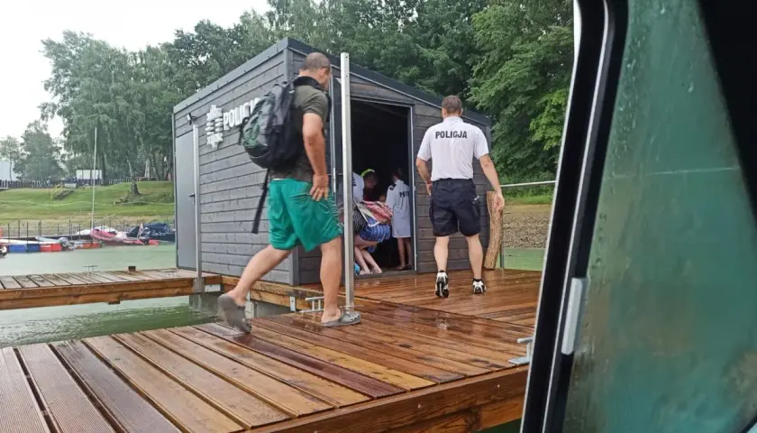 Policjanci na pomoście nad Soliną, przygotowani na nagłe zmiany pogody. Widać budynek z napisem "POLICJA" i łodzie w tle.